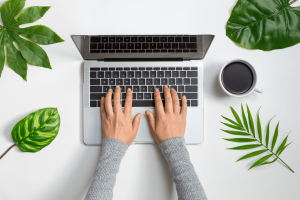 A person typing laptop computer surrounded by plants and a coffee mug