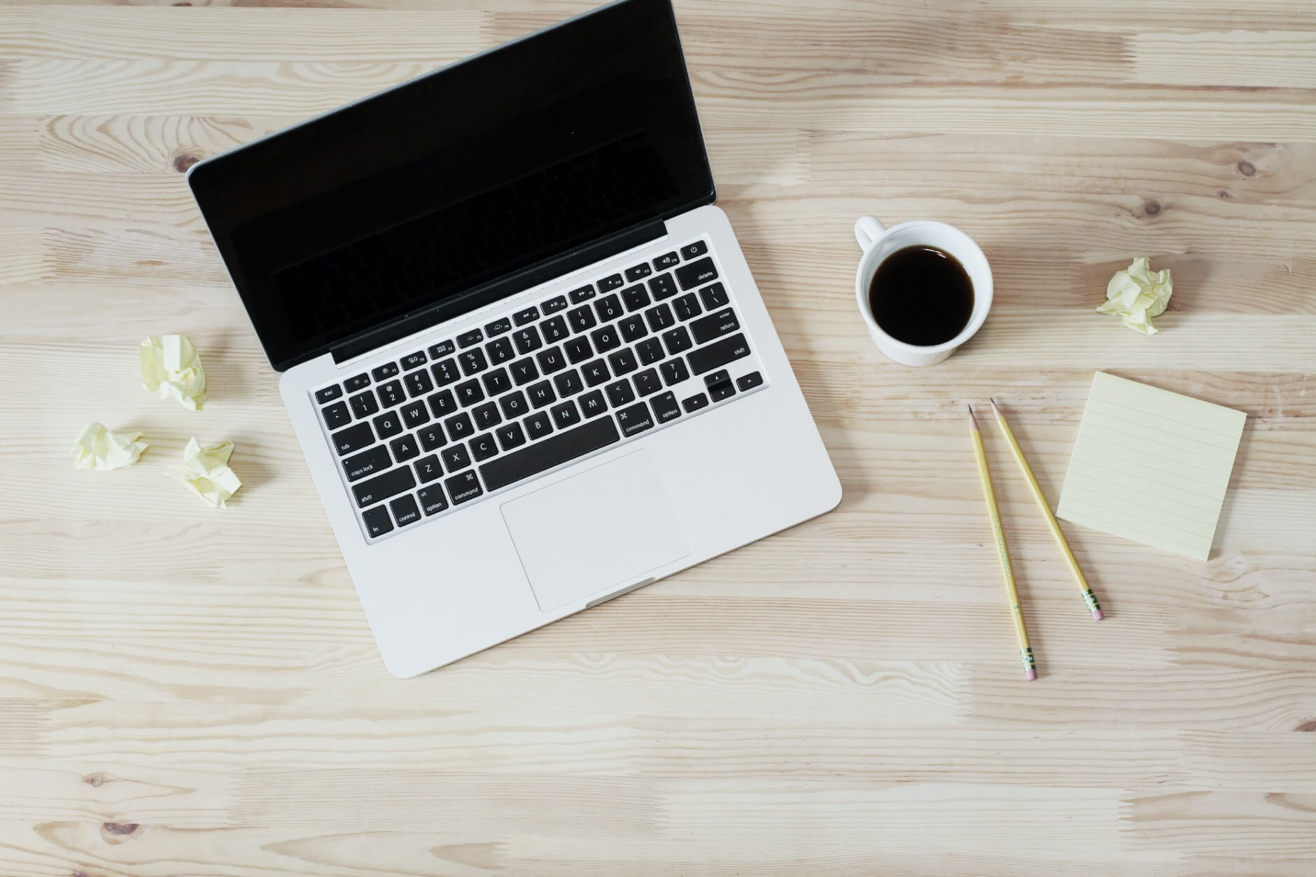 a laptop with crumpled balls of paper, cup of coffee, pencils, and notepad